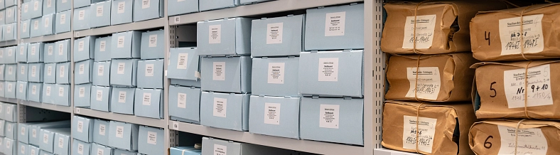 Blick in ein Archivregal. Links hellblaue Archivkartons, rechts Aktenpakete in braunes Packpapier eingeschlagen.